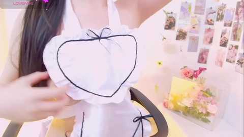 Media: Video of an Asian woman with long black hair, wearing a revealing white maid outfit with a heart-shaped chest piece, posing in a bright, pastel-colored room with floral decorations and photos on the wall.