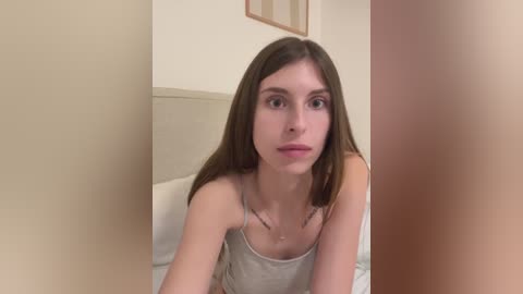 Media: Video of a young, slender, fair-skinned woman with straight, shoulder-length brown hair, wearing a light gray tank top, sitting on a bed with white sheets in a beige room.