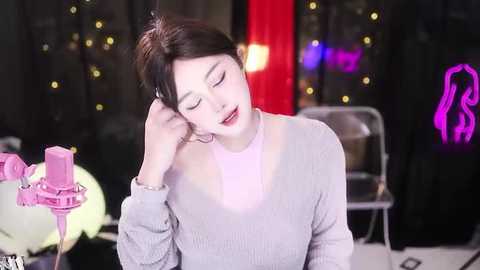 Media: Video of a young woman with light skin, wearing a light pink sweater, resting her head on her hand, in a dimly lit room with glowing strings of lights and a neon sign in the background.