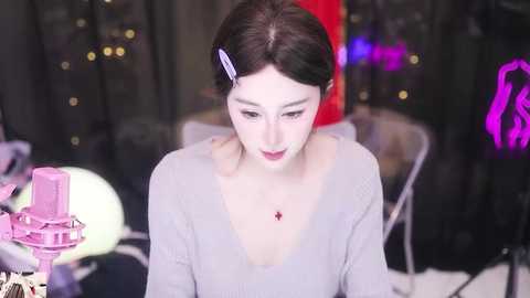 Media: Video of a young Asian woman with fair skin, dark hair, and a purple hair clip, wearing a light grey sweater, standing in a dimly lit room with blurred backgrounds.