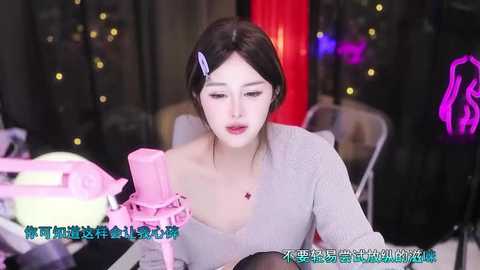 Media: A video of a young Asian woman with fair skin, dark hair, and a blue hairpin, wearing a light grey off-shoulder top, sitting at a makeup table with pink tools, in a dimly-lit room with black curtains and glowing lights.