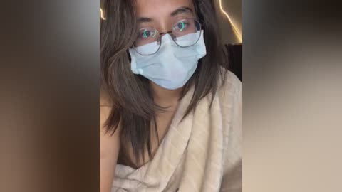 Media: Video of a young woman with long, dark hair and green eyes, wearing a light blue medical mask and rectangular glasses, wrapped in a beige towel, against a blurred indoor background.