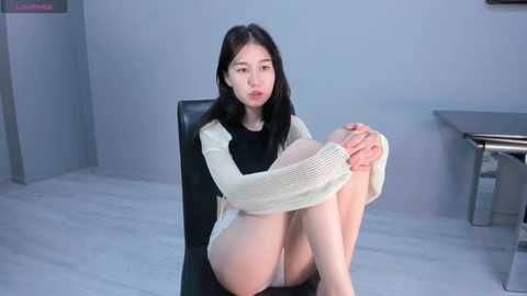 Media: A video of an East Asian woman with fair skin, black hair, and a slender physique, wearing a white knitted sweater and white panties, sitting on a black chair in a modern, minimalist room with light-colored walls and a glass table.