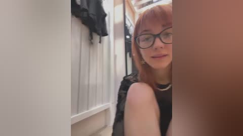 Media: Video of a young woman with light skin and shoulder-length red hair, wearing black-rimmed glasses, a black sleeveless top, and a silver necklace. She is partially hidden by a wooden door frame in a narrow hallway.