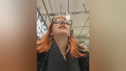 Media: A close-up video of a woman with red hair, wearing glasses, and a black jacket, standing in a modern, brightly-lit room with exposed ceiling beams and white walls.