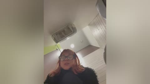 Media: Video of a woman with red hair, wearing black-framed glasses and a black fur coat, standing in a modern, brightly lit restroom with white walls, a glass partition, and a green accent.