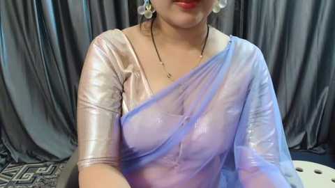 Media: Video of a light-skinned woman with medium-length dark hair, wearing a shiny, translucent silver saree with a blue drape, and white pearl earrings, against a gray, patterned backdrop.