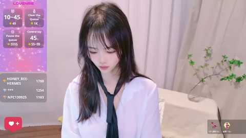 Media: A video of an Asian woman with long black hair, wearing a white shirt and black tie, standing in a bedroom with a white bed and plant.