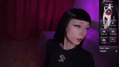 Video of a pale-skinned woman with straight black hair, wearing black makeup, black dress, and sitting against a red curtain background. She appears to be in a dimly lit room, with a digital screen displaying her stats.