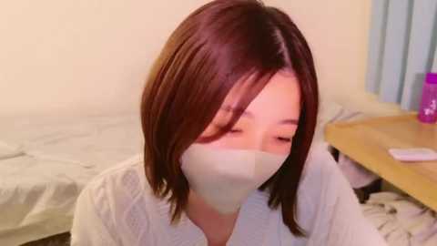 Media: Video of an Asian woman with shoulder-length brown hair, wearing a white mask, sitting on a bed in a dimly lit room. Background includes a wooden nightstand with a pink object and a bed with white sheets.