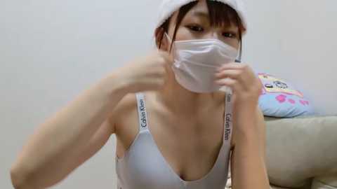 Media: A young Asian woman with a slender physique, wearing a white Calvin Klein bra and mask, sits indoors, holding a white towel, with a pink and blue pillow in the background.