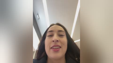 Video of a young woman with fair skin, long dark hair, and a slightly open mouth, captured from a low angle, in an indoor setting with bright, recessed ceiling lights.