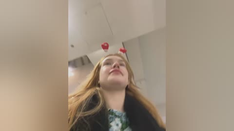 Media: A video of a young woman with long, straight blonde hair, wearing a green floral top, standing in a dimly lit room. Two red heart-shaped stickers are attached to her forehead, giving a playful, whimsical touch to the image.