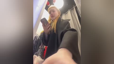 Media: A video captures a blonde woman in a black coat, texting, in a dimly lit, cramped changing room filled with clothes.