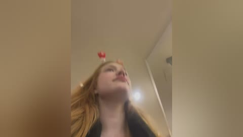 Media: Video of a young woman with long, straight blonde hair wearing a black top, standing in a beige room. She has a red heart-shaped sticker on her forehead, looking up.