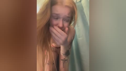 Media: A blurry video of a naked, fair-skinned, red-haired woman with tattoos, covering her face with her hands in a bathroom with a shower curtain.