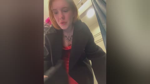 Media: Video of a fair-skinned, red-haired woman with a black blazer over a red top, wearing a black necklace. She is leaning forward, looking down. Background features a blue curtain and a white door.