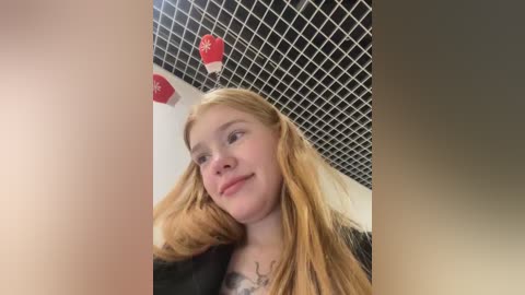 Media: Video of a young Caucasian woman with long, wavy blonde hair, wearing a black top, standing indoors with a grid-patterned ceiling. She has a neutral expression, and red and white balloons float above her.