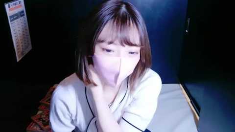 Media: A video shows an Asian woman with shoulder-length brown hair, wearing a white blouse, hiding her mouth with her hands, against a dark, dimly lit background.