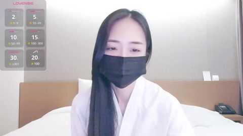 Media: Video of a woman with long black hair and light skin, wearing a white robe and black face mask, sitting on a bed in a minimalist room with a wooden headboard and digital thermometer displaying 15\u00b0C.