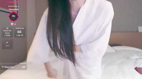 Media: Video of a woman with long black hair in a white robe, standing in a modern bedroom with a bed and wooden headboard.