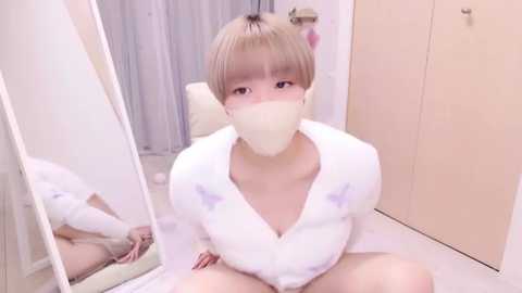 Media: Video of a young Asian woman with short, platinum blonde hair, wearing a white bathrobe and face mask, sitting on a tiled floor in a minimalist bathroom with beige cabinets and a mirror.