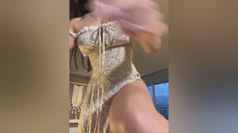 Media: Video of a woman with long dark hair, wearing a floral off-shoulder dress, captured mid-motion, in a dimly lit room with a window and hanging decor in the background.