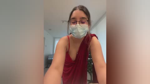 Media: Video of a young woman with light skin, brown hair, wearing a red sleeveless top, a blue surgical mask, and rectangular glasses. She is standing in a hallway with white walls and a potted plant in the background.