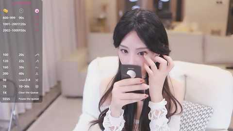 Media: A video of an Asian woman with long black hair, wearing a white lace top, holding a black smartphone. She is in a modern, minimalist room with soft, neutral tones.