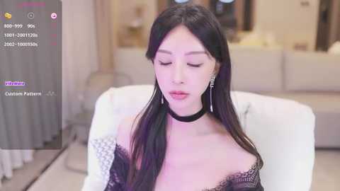 Media: Video of an Asian woman with long black hair, wearing black lace lingerie and a choker, seated on a white chair in a modern, softly lit room.