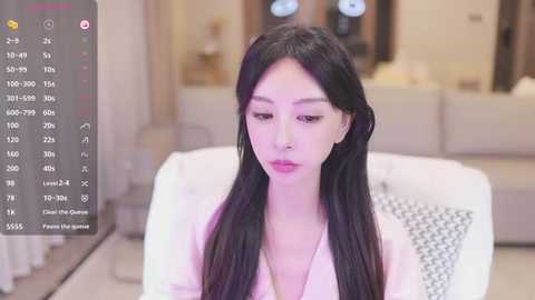 Media: A video of an East Asian woman with long black hair, light skin, and pink lipstick, wearing a light pink top, sitting indoors with a blurred background.