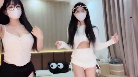 Media: Video of two Asian women with long black hair, wearing white crop tops, white shorts, and face masks, in a modern bedroom with beige walls and minimalistic decor.