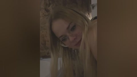 Media: Video of a young, fair-skinned woman with long blonde hair, wearing glasses, leaning forward, smiling, with a dimly lit bathroom background.