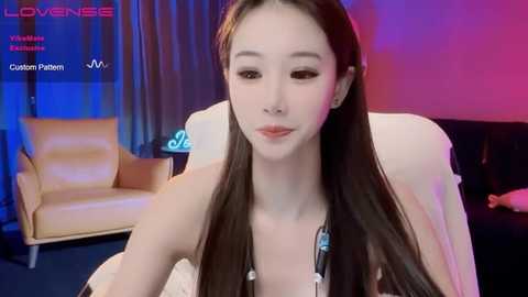 Media: Video of a slender East Asian woman with long, straight brown hair, wearing a white top, seated indoors. Background includes a modern armchair, blue and pink lighting, and a \"Lovense\" watermark.