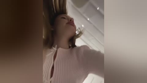 Media: Video of a woman with fair skin and brown hair, wearing a white ribbed knit top, lying on her back with eyes closed, surrounded by soft, white, and beige background lighting.