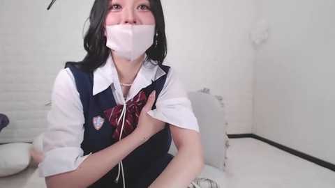 Media: A video of an Asian woman with long black hair, wearing a white shirt, blue vest, and red tie, gagged, sitting on a white toilet, in a white-tiled bathroom.