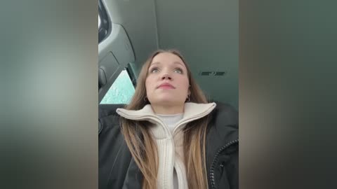 Media: Video of a young Caucasian woman with long brown hair, wearing a black jacket and beige scarf, sitting in an airplane seat, gazing out the window.