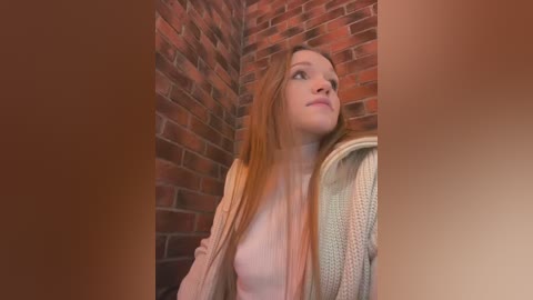 Media: A video of a young Caucasian woman with long, straight, light brown hair, wearing a light pink top and a beige cardigan, standing in front of a brick wall.