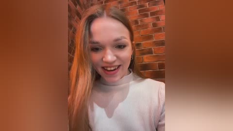 Media: Video of a smiling, fair-skinned woman with long blonde hair, wearing a white, ribbed sweater, standing against a red brick wall.