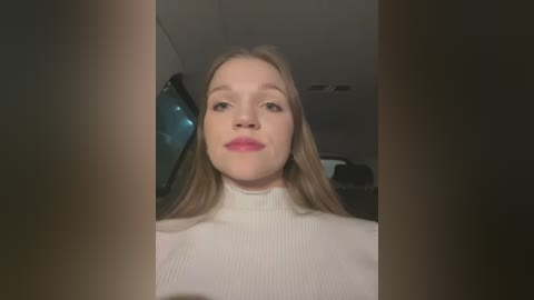 Media: Video of a young Caucasian woman with long blonde hair, fair skin, and a neutral expression, wearing a white turtleneck sweater, standing indoors with a blurry background.