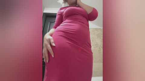 Media: A video of a pregnant woman in a tight, bright pink dress, standing in a dimly lit room with beige tiles and a black door in the background.