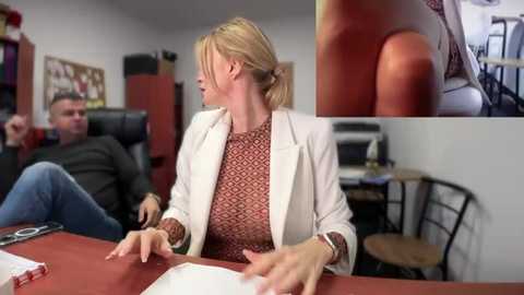 Media: Video of a blonde woman in a red-patterned blouse, white blazer, and tan wristwatch, seated at a red desk, speaking to a man in a gray sweater and blue jeans in an office with red chairs, wooden stools, and shelves.