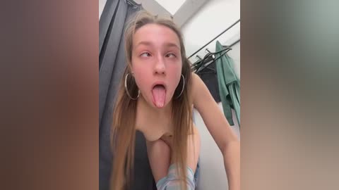 Media: A video of a young Caucasian woman with long blonde hair, wearing a plaid top, sticking out her tongue playfully in a mirrored bathroom with clothes hanging on a rack.