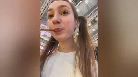 Media: Video of a young woman with light skin and long brown hair in pigtails, wearing a white shirt, eating a popsicle in a brightly lit, modern indoor setting with visible ceiling beams.