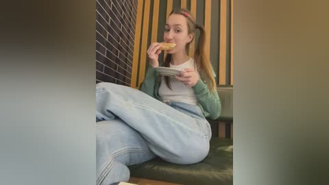 Media: Video of a young woman with light skin, long brown hair, and pigtails, wearing a green sweater and light blue jeans, eating a sandwich on a dark green couch against a brick and vertical-striped wall.