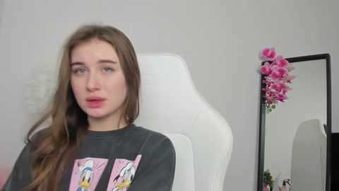 Media: Video of a young woman with long brown hair, wearing a dark graphic T-shirt, seated in a white gaming chair. The background features a wall with a pink flower arrangement and a large mirror.