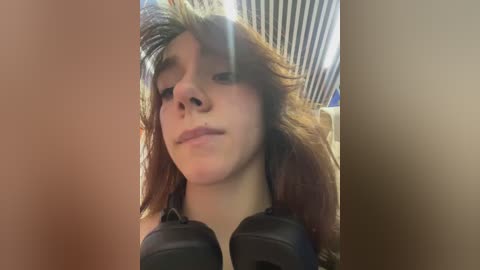 Media: Video of a young person with long, tousled brown hair wearing black headphones, slightly blurry, taken indoors with vertical striped wallpaper in the background.