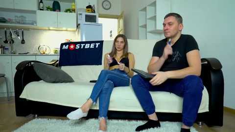 Media: Video of a man and woman in casual clothes, sitting on a beige sofa in a modern kitchen with white cabinets and a blue \"MOSTBET\" banner.