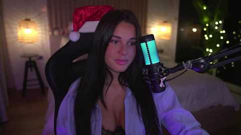 Media: Video of a young woman with long black hair, wearing a red Santa hat, a light shirt, and a black bra, singing into a microphone in a dimly-lit bedroom with Christmas lights.