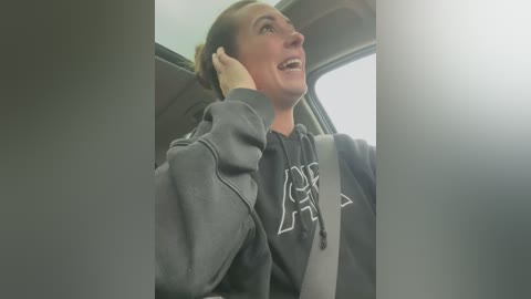 Media: Video of a woman with short brown hair, wearing a gray hoodie with a white logo, smiling while holding her ear, seated in a car with a gray seatbelt.